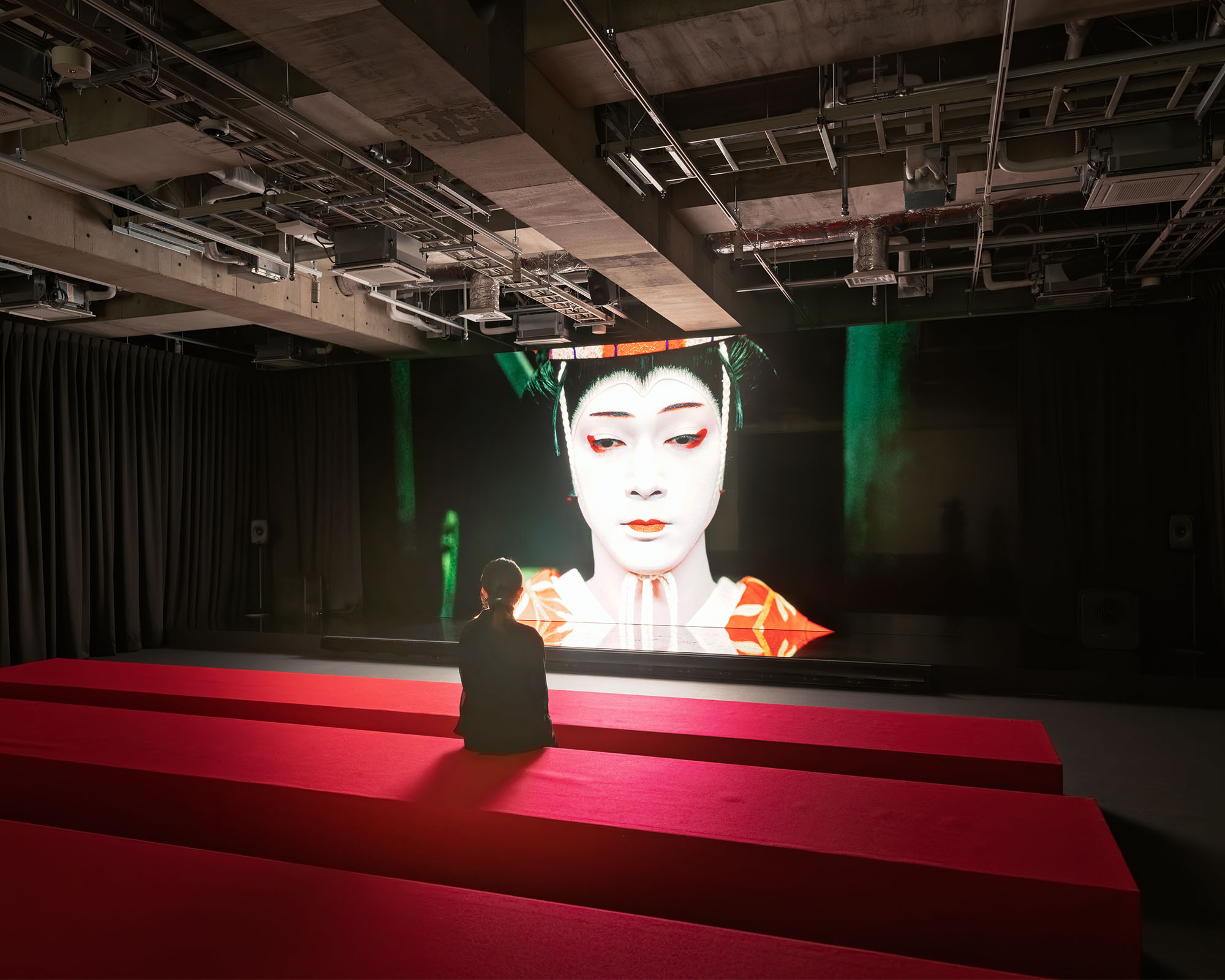 The theme song 'Luminance' and the main footage are screened in an exhibition room with dimmed lighting. In front of the screen, a red bench has been provided, creating a space where visitors can sit and enjoy the viewing.