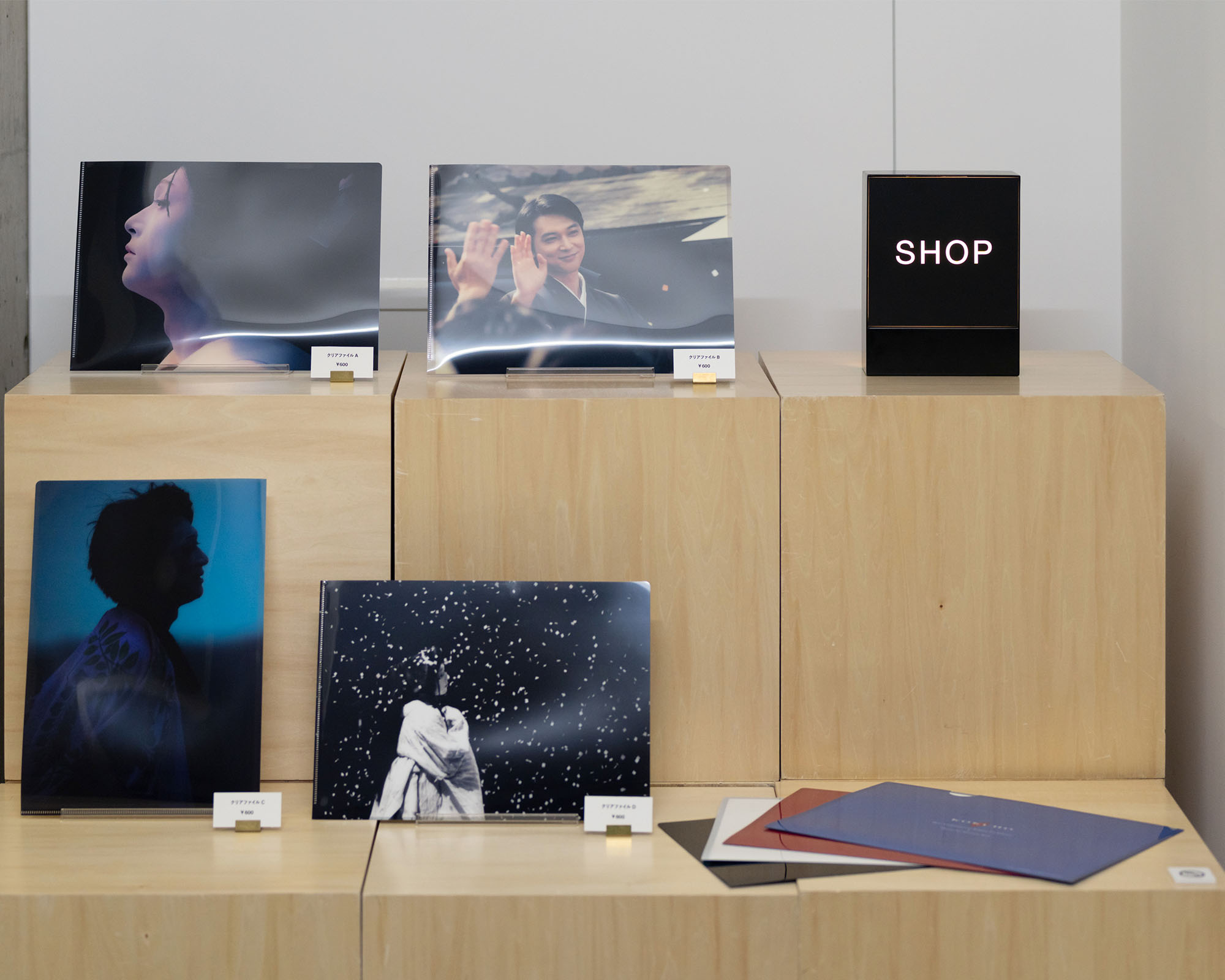 Exhibition-related items displayed at the shop on 4F, related merchandise arranged on wooden stands. The items are neatly arranged on a wooden platform.