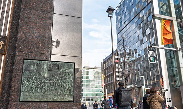 銀座二丁目交差点の大倉本館角に埋め込まれた「東京銀座通電気燈建設之図」のレリーフ。通り沿いには復刻したアーク灯が建てられている。